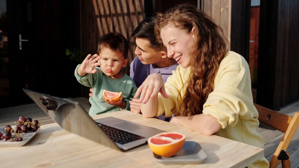A family sitting outside enjoying a video call on a laptop, sharing smiles and fresh fruit.